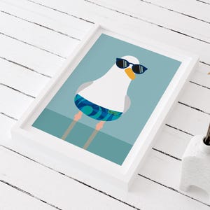 May include: Framed art print of a cartoon seagull wearing sunglasses and swim shorts. The seagull has a white body, grey wings, and an orange beak. The background is a light blue colour.