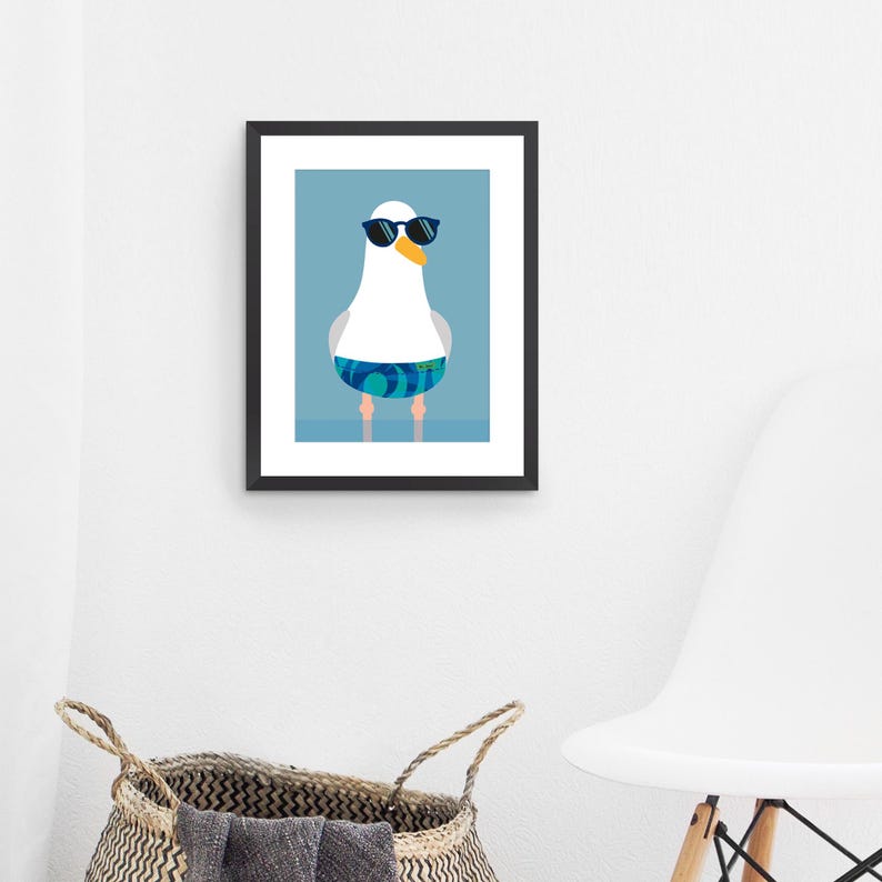 May include: A framed art print of a cartoon seagull wearing sunglasses and swim shorts. The seagull is white with a yellow beak and blue and green patterned swim shorts. The background is light blue.