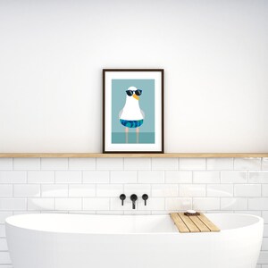 May include: A framed art print depicting a cartoon seagull wearing sunglasses and swimming trunks. The seagull is white with blue and green patterned trunks. The background is a light blue colour.