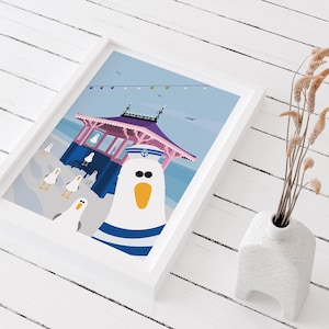 May include: A white seagull wearing a blue and white sailor hat stands in front of a pink and blue beach hut with a blue roof. The seagull has a yellow beak and black eyes. There are other seagulls in the background.