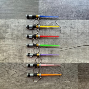 May include: Eight lightsaber keychains with different colored blades. The blades are blue, yellow, red, green, purple, pink, orange, and black. The keychains are all silver and black.