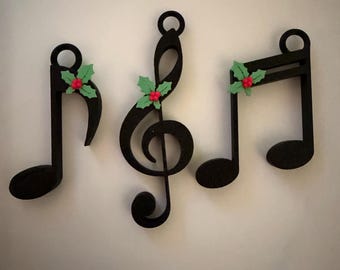 Könnte beinhalten: Drei schwarze Notenornamente mit Stechpalmenakzenten. Die Ornamente umfassen eine Achtelnote, einen Violinschlüssel und eine halbe Note. Jedes Ornament hat grüne Stechpalmenblätter und rote Beeren für die Weihnachtsdekoration.