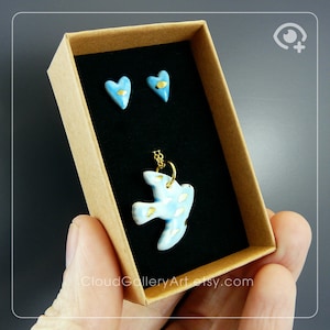 Könnte beinhalten: Ein Schmuckset mit einer hellblauen Vogel-Anhänger-Halskette und passenden herzförmigen Ohrsteckern. Der Vogel und die Herzen haben goldfarbene Akzente. Das Set wird in einer braunen Schachtel mit schwarzem Innenraum präsentiert.
