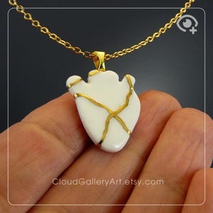 Puede incluir: Un colgante blanco con detalles dorados, suspendido de una cadena dorada. El colgante tiene una forma única con líneas doradas, posiblemente un diseño Kintsugi. El collar se muestra sobre un fondo oscuro.