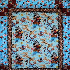 May include: A blue and brown patchwork quilt with a pirate theme. The center panel features a repeating pattern of a pirate ship, a treasure chest, and a map with the words "Pirate Island" and "Dead Sea".