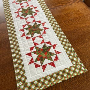 May include: A quilted Christmas table runner featuring a central design of red and green stars on a white background. The stars are filled with festive patterns. The runner is bordered with a green and red plaid fabric.