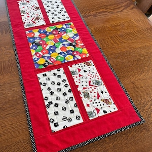 May include: A rectangular red table runner with a black and white polka dot border. The center features fabric panels with playing cards, dice, and a colorful pool ball design. The runner is placed on a wooden table.
