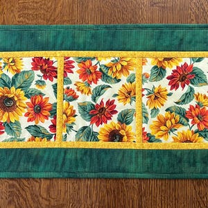 May include: Rectangular table runner featuring a floral pattern of sunflowers and red flowers on a cream background. The center design is framed by a yellow border and a green plaid border.
