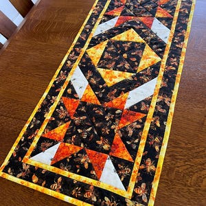 May include: A rectangular table runner with a bee-themed design. The runner features a patchwork pattern with black, orange, white, and yellow geometric shapes. The fabric is decorated with bee illustrations, creating a warm and inviting aesthetic.