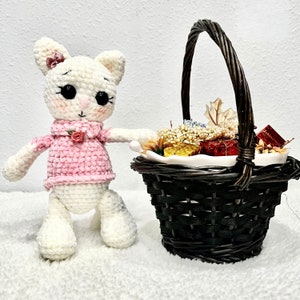 May include: A white crocheted cat wearing a pink sweater with a small pink flower on the collar. The cat is standing next to a brown wicker basket filled with decorative items.