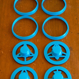 May include: Eight turquoise plastic cookie cutters in various shapes. There are four circular cutters and four cutters with a central knob and decorative cutouts. The cutters are arranged on a wooden surface.