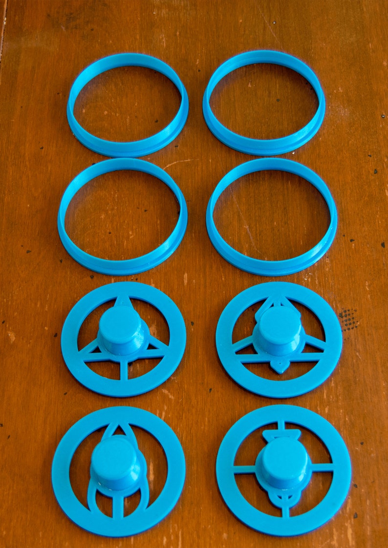 May include: Eight turquoise plastic cookie cutters in various shapes. There are four circular cutters and four cutters with a central knob and decorative cutouts. The cutters are arranged on a wooden surface.