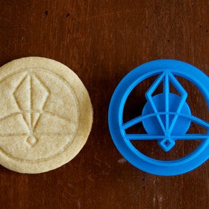 May include: A round, light-colored cookie with an embossed geometric design next to a blue plastic cookie cutter. The cookie cutter is in the shape of the design on the cookie. The items are on a dark brown wooden surface.