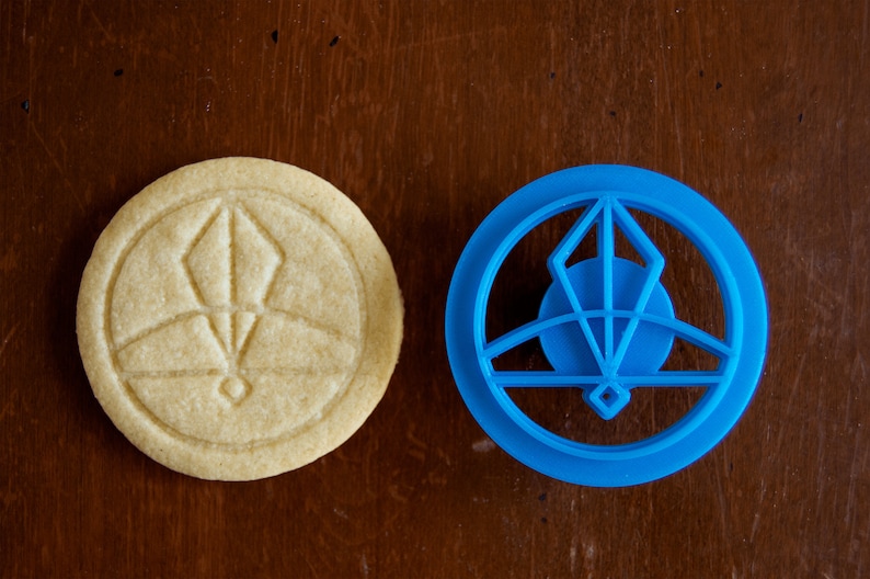 May include: A round, light-colored cookie with an embossed geometric design next to a blue plastic cookie cutter. The cookie cutter is in the shape of the design on the cookie. The items are on a dark brown wooden surface.