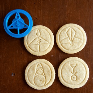 May include: A blue plastic cookie cutter and four round cookies with embossed geometric designs. The cookies are light brown and have various symbols. The cookie cutter is in the shape of a circle with a triangle and other shapes inside.