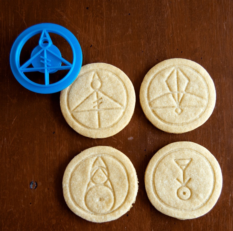 May include: A blue plastic cookie cutter and four round cookies with embossed geometric designs. The cookies are light brown and have various symbols. The cookie cutter is in the shape of a circle with a triangle and other shapes inside.