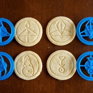 May include: An overhead shot shows six round cookies with embossed designs and two blue cookie cutters. The cookies are beige, and the cutters are blue. The designs include geometric shapes and symbols. The cookies and cutters are on a brown surface.