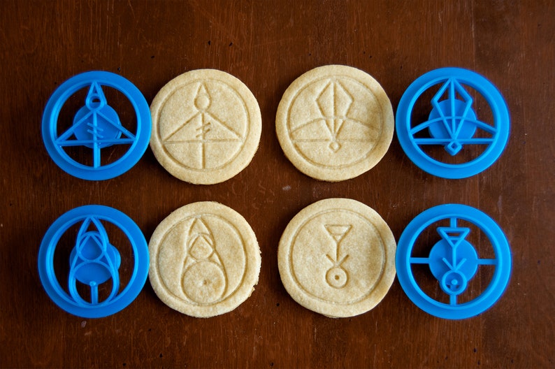 May include: An overhead shot shows six round cookies with embossed designs and two blue cookie cutters. The cookies are beige, and the cutters are blue. The designs include geometric shapes and symbols. The cookies and cutters are on a brown surface.