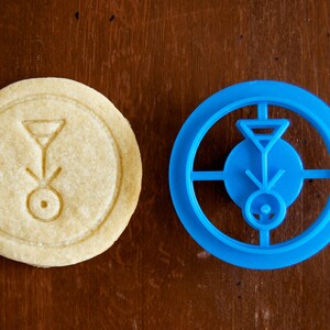 May include: A round, light-colored cookie with an embossed design next to a blue cookie cutter. The cookie has a circular shape with a geometric symbol in the center. The blue cutter mirrors the design.