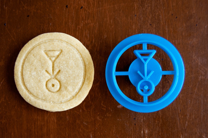 May include: A round, light-colored cookie with an embossed design next to a blue cookie cutter. The cookie has a circular shape with a geometric symbol in the center. The blue cutter mirrors the design.