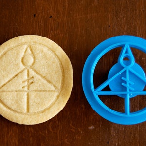 May include: A light-colored, round cookie with an embossed design next to a blue plastic cookie cutter. The cookie has a circular border with a triangle and vertical lines inside. The cookie cutter is the same shape.