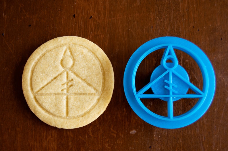 May include: A light-colored, round cookie with an embossed design next to a blue plastic cookie cutter. The cookie has a circular border with a triangle and vertical lines inside. The cookie cutter is the same shape.