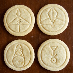 May include: Four round, light-colored cookies with intricate embossed designs. Each cookie features a different geometric symbol within a circular border. The cookies are arranged on a dark brown wooden surface, creating a visually appealing contrast.