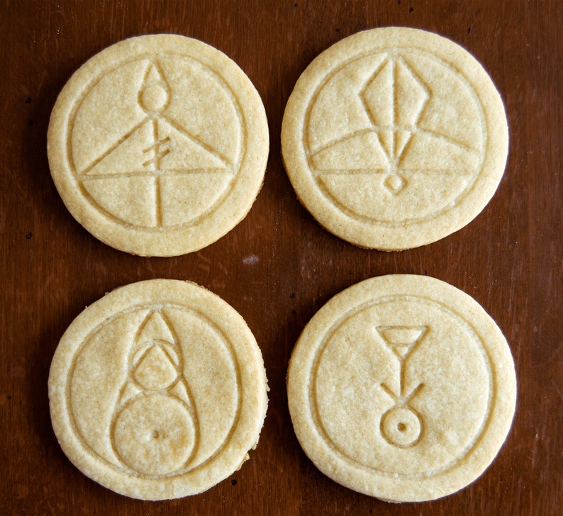May include: Four round, light-colored cookies with intricate embossed designs. Each cookie features a different geometric symbol within a circular border. The cookies are arranged on a dark brown wooden surface, creating a visually appealing contrast.