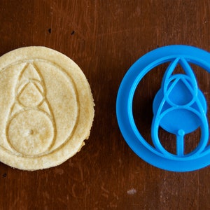 May include: A round, baked cookie with a detailed design next to a blue plastic cookie cutter. The cookie has a light brown color and the cutter is a vibrant blue. The design on both is a stylized shape within a circle.