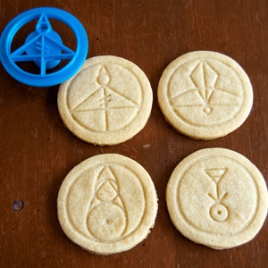 May include: A blue plastic cookie cutter and four round cookies with embossed geometric designs. The cookies are light brown and sit on a dark brown wooden surface. The cookie cutter is in the upper left corner.