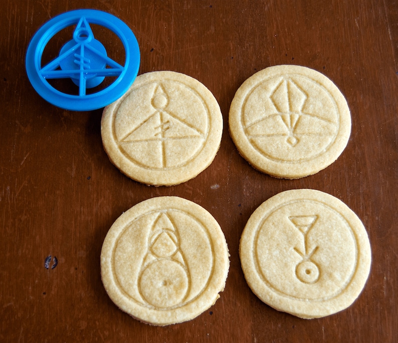 May include: A blue plastic cookie cutter and four round cookies with embossed geometric designs. The cookies are light brown and sit on a dark brown wooden surface. The cookie cutter is in the upper left corner.