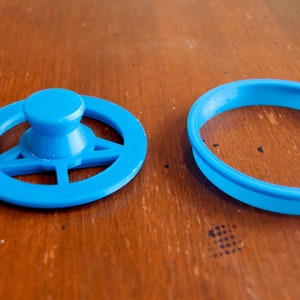 May include: Two bright blue plastic cookie cutters. One is a steering wheel shape with a central knob, and the other is a simple round shape. The cutters are on a wooden surface.