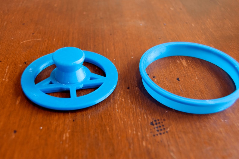May include: Two bright blue plastic cookie cutters. One is a steering wheel shape with a central knob, and the other is a simple round shape. The cutters are on a wooden surface.