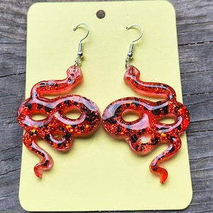 May include: A pair of red snake-shaped earrings with silver-colored hooks. The earrings are made of a translucent red material with black and gold glitter embedded throughout. They are displayed on a yellow card.