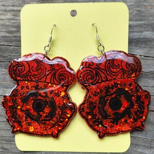 Puede incluir: Un par de pendientes rojos con purpurina con un diseño de remolino negro y una forma negra central. Los pendientes están sobre una tarjeta amarilla con un agujero en la parte superior. Los pendientes tienen ganchos plateados.
