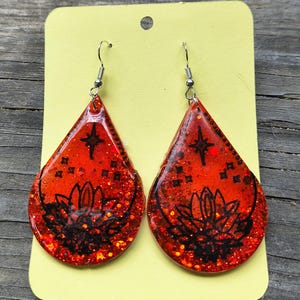 May include: A pair of teardrop-shaped earrings with a red glitter background. Each earring features a black lotus flower design and a star. The earrings have silver-colored ear wires.