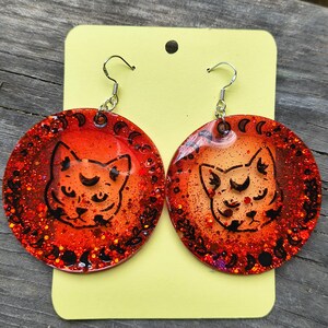 Puede incluir: Un par de pendientes redondos, naranja y rojo con purpurina. Cada pendiente presenta una cara de gato negro con una luna creciente y un borde de lunas crecientes y hojas. Los pendientes cuelgan de ganchos plateados.
