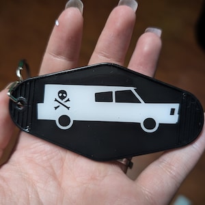 May include: Black plastic keychain shaped like a motel key with a white silhouette of a hearse with a skull and crossbones on the side.