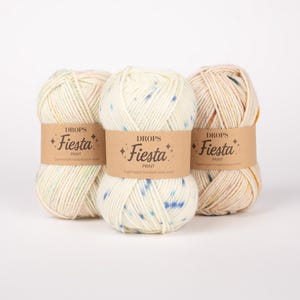 May include: Three skeins of Drops Fiesta Print yarn, each wrapped in a tan paper label with the text Drops Fiesta Print. The yarn is an off-white color with small, scattered flecks of blue, orange, and yellow throughout the textured fibers.