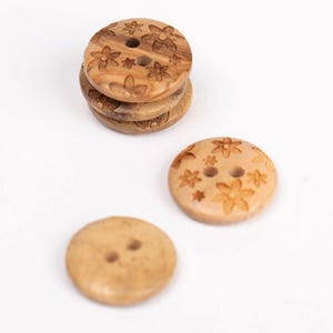 May include: A set of four wooden buttons with a floral design. The buttons are round and have two holes. The buttons are a light brown color.