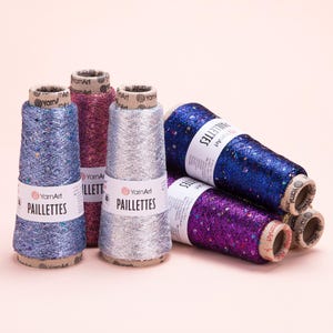 May include: Five spools of YarnArt Paillettes yarn in blue, pink, silver, dark blue, and purple. Each spool features a metallic, shimmering texture with embedded sequins and a white paper label with the brand name and product text clearly visible.