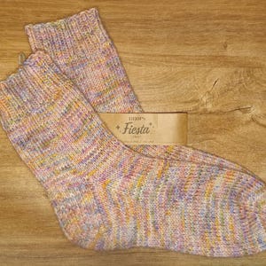 May include: A pair of hand-knitted socks featuring a variegated yarn pattern with soft pastel shades of pink, yellow, blue, and purple. A tan paper label is attached to the socks, displaying the text Drops Fiesta in a decorative font.