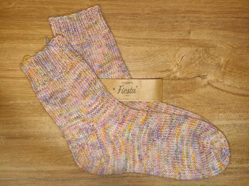 May include: A pair of hand-knitted socks featuring a variegated yarn pattern with soft pastel shades of pink, yellow, blue, and purple. A tan paper label is attached to the socks, displaying the text Drops Fiesta in a decorative font.
