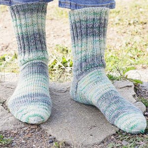 May include: A pair of mid-calf knit socks featuring a variegated color pattern in shades of green, blue, and cream. The socks are constructed with a ribbed texture throughout the leg and foot, ending in a rounded toe. The design includes a visible heel.