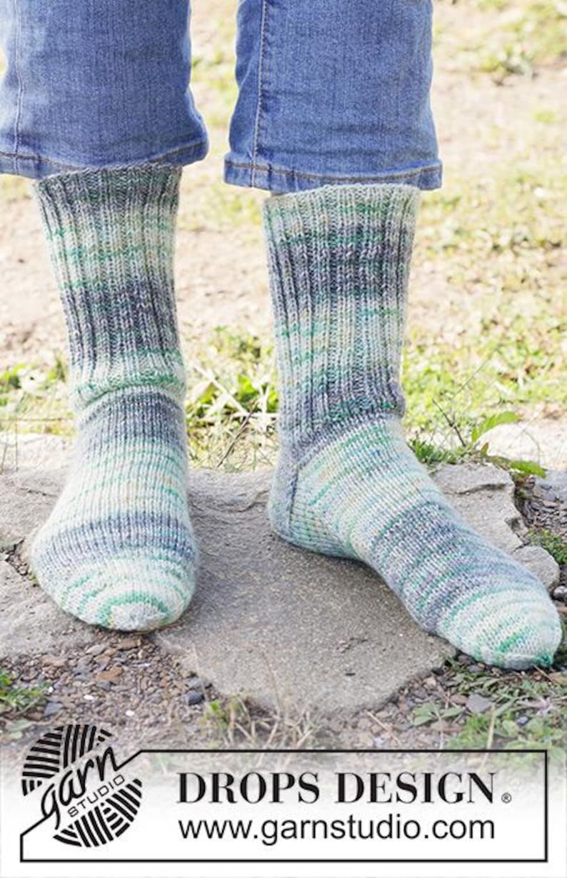 May include: A pair of mid-calf knit socks featuring a variegated color pattern in shades of green, blue, and cream. The socks are constructed with a ribbed texture throughout the leg and foot, ending in a rounded toe. The design includes a visible heel.