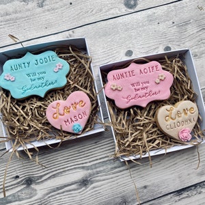 Puede incluir: Dos cajas de regalo blancas con papel triturado marr&oacute;n en su interior. Cada caja contiene dos galletas decoradas. Una caja tiene una galleta azul claro con el texto "AUNTY JODIE Will you be my Godmother?" y una galleta en forma de coraz&oacute;n rosa con el texto "Love MASON". La otra caja tiene una galleta rosa con el texto "AUNTIE AOIFE Will you be my Godmother?" y una galleta en forma de coraz&oacute;n beige con el texto "Love CLIODHNA".