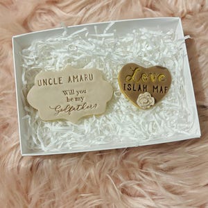 Puede incluir: Una caja blanca con papel de seda blanco en su interior. La caja contiene dos peque&ntilde;as galletas decoradas. Una galleta tiene forma de coraz&oacute;n y dice "Love Islah Maf" en letras doradas. La otra galleta es rectangular y dice "Uncle Amaru Will you be my Godfather?" en letras doradas.