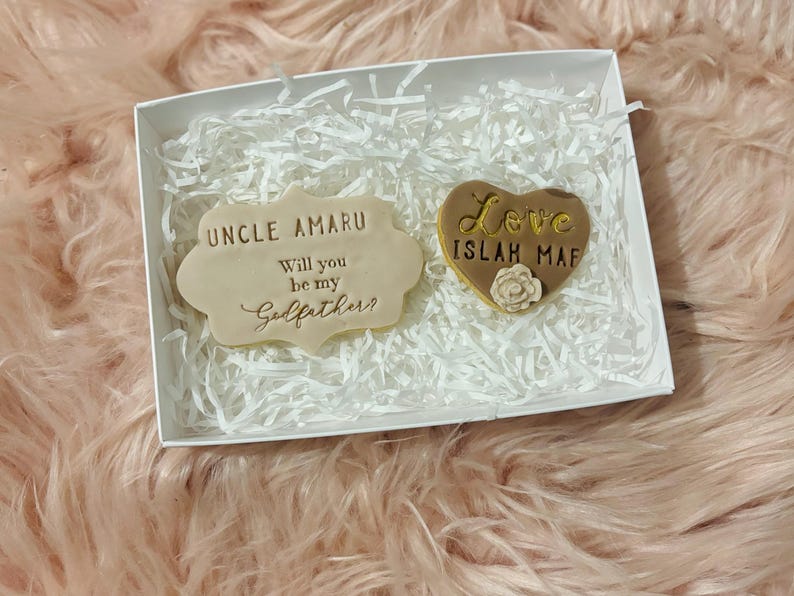 Puede incluir: Una caja blanca con papel de seda blanco en su interior. La caja contiene dos peque&ntilde;as galletas decoradas. Una galleta tiene forma de coraz&oacute;n y dice "Love Islah Maf" en letras doradas. La otra galleta es rectangular y dice "Uncle Amaru Will you be my Godfather?" en letras doradas.