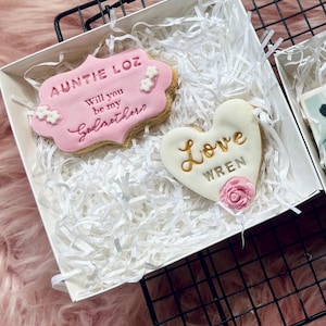 Puede incluir: Una caja blanca con dos galletas. Una galleta tiene forma de coraz&oacute;n y dice "Love Wren" en letras doradas. La otra galleta es rectangular y dice "Auntie Loz Will you be my Godbrother?" en letras rosas y doradas.