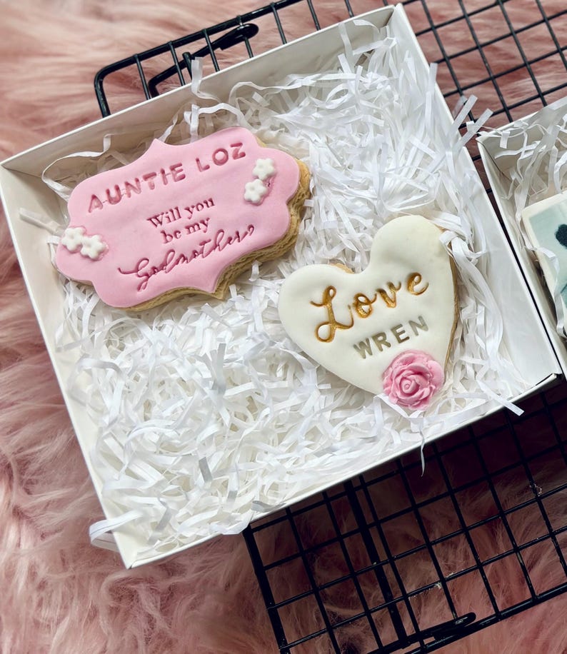 Puede incluir: Una caja blanca con dos galletas. Una galleta tiene forma de coraz&oacute;n y dice "Love Wren" en letras doradas. La otra galleta es rectangular y dice "Auntie Loz Will you be my Godbrother?" en letras rosas y doradas.